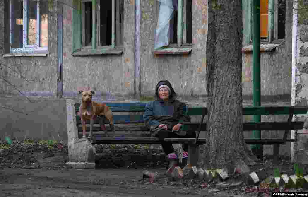 Жінка із собакою сидить на лавці в Часовому Яру. У будинку вибиті усі вікна. 9 квітня 2023 року. Часів Яр, який розташований на височині лише за кілька кілометрів на захід від Бахмута, ймовірно, стане наступним оплотом ЗСУ проти російських сил. Українське командування стверджує, що російські сили у Бахмуті застосовують сирійську тактику «випаленої землі», що знищує все до тла.