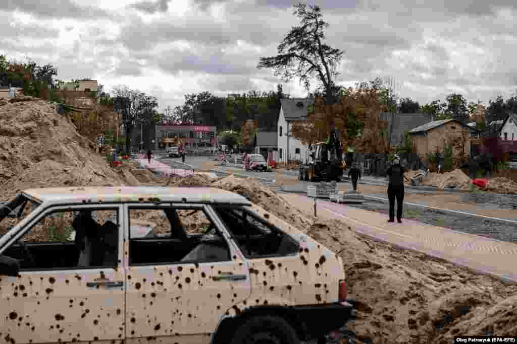 Muncitorii din Irpin, un oraș de la periferia capitalei ucrainene Kiev, reconstruiesc un drum distrus în bombardamentele rusești masive din martie 2022. Fotografia e făcută pe 23 octombrie 2023. Mașina ciuruită este acum o amintire sumbră a luptei crâncene de anul trecut.